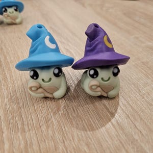 May include: Two charming frog figurines, each wearing a wizard's hat. One hat is blue with a white crescent moon, the other purple with a yellow crescent moon. The frogs are light green and have smiling faces.