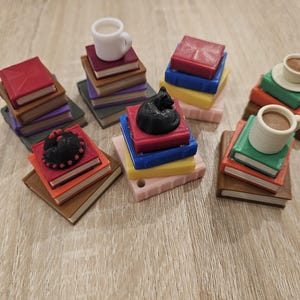 May include: Miniature stacks of books in various colours, topped with a white mug, a black cat figurine, and a decorative black circle. The books are brown, red, green, yellow, and pink. The mugs contain a brown liquid.