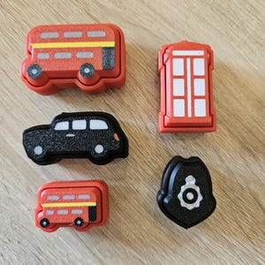 May include: A set of toy London-themed items: a red double-decker bus, a black car, a red telephone box, and a police badge. The toys are brightly coloured and designed in a simple, cartoon style, perfect for children.