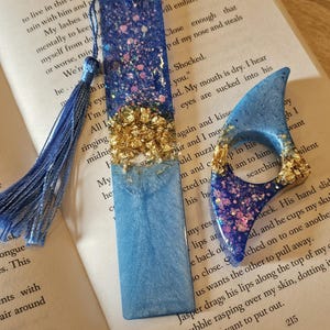 May include: A blue resin bookmark and a matching thumb rest. The bookmark has a blue tassel and is decorated with gold leaf and glitter. The thumb rest is also blue with gold leaf and glitter accents. Both are on a book.