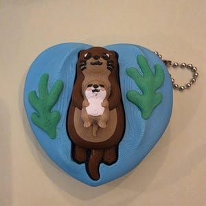 May include: A brown otter holding a smaller otter, set against a blue heart-shaped background with green seaweed accents. The keychain charm has a silver chain and is made of a textured material.