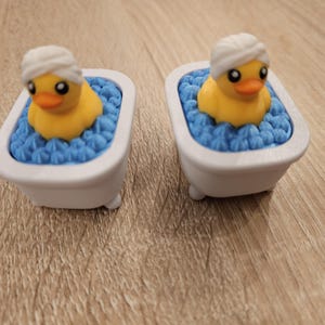 May include: Two miniature bathtubs, each with a yellow rubber duck wearing a white towel. The ducks are in white bathtubs filled with blue foam. The bathtubs are on a light brown wooden surface.
