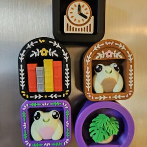 May include: A selection of decorative magnets with diverse designs. Includes a clock, a bookshelf with colourful books, a frog, and a potted plant. The magnets are in black, brown, purple, and white.