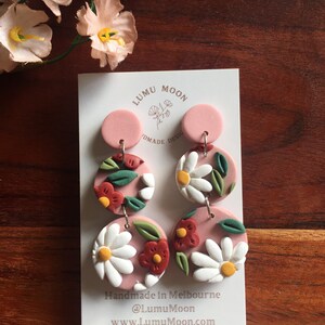 May include: A pair of pink and white floral dangle earrings with a pink circle at the top. The earrings are handmade in Melbourne, Australia. The text "Handmade in Melbourne @LumuMoon www.LumuMoon.com" is printed on the card.