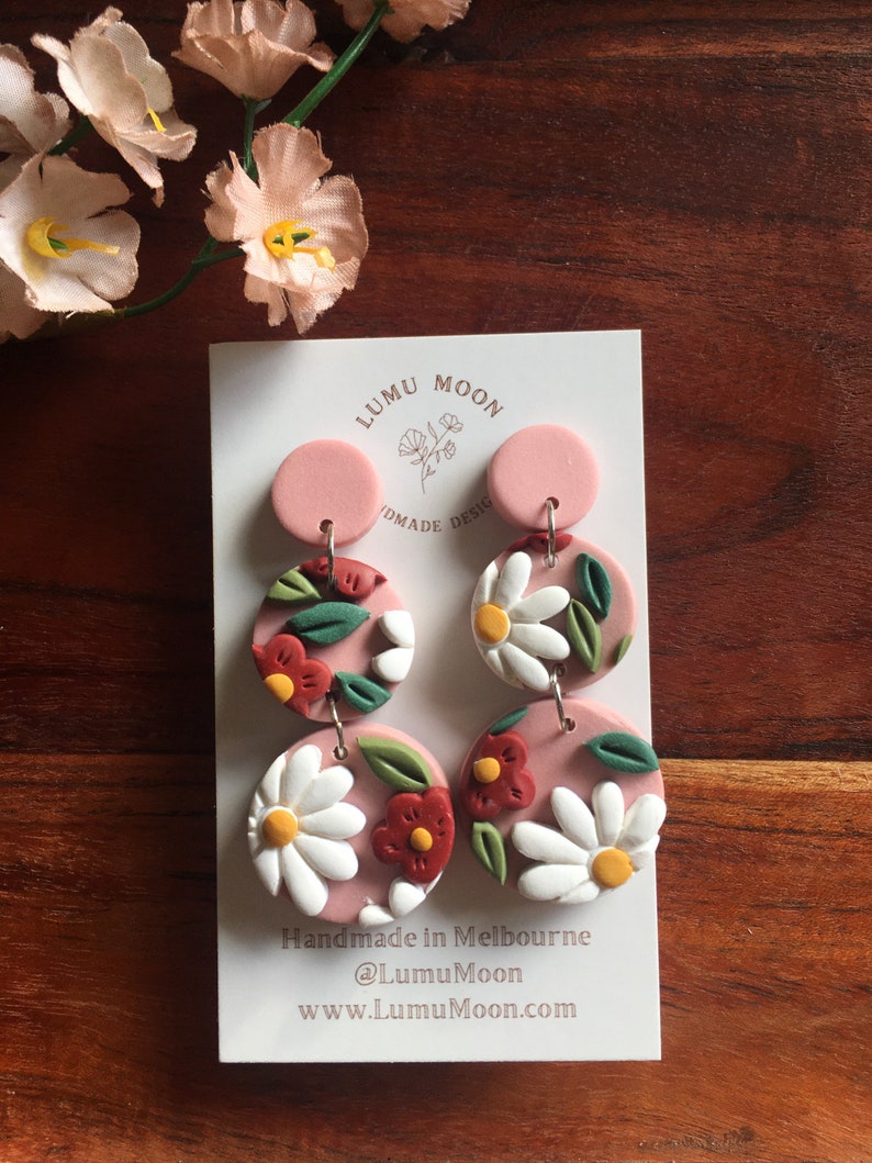 May include: A pair of pink and white floral dangle earrings with a pink circle at the top. The earrings are handmade in Melbourne, Australia. The text "Handmade in Melbourne @LumuMoon www.LumuMoon.com" is printed on the card.