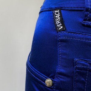 May include: Close-up of a pair of royal blue pants. The pants feature a small pocket, a silver button, and the "VERSACE" label. The fabric has a satin sheen, and the stitching is visible.