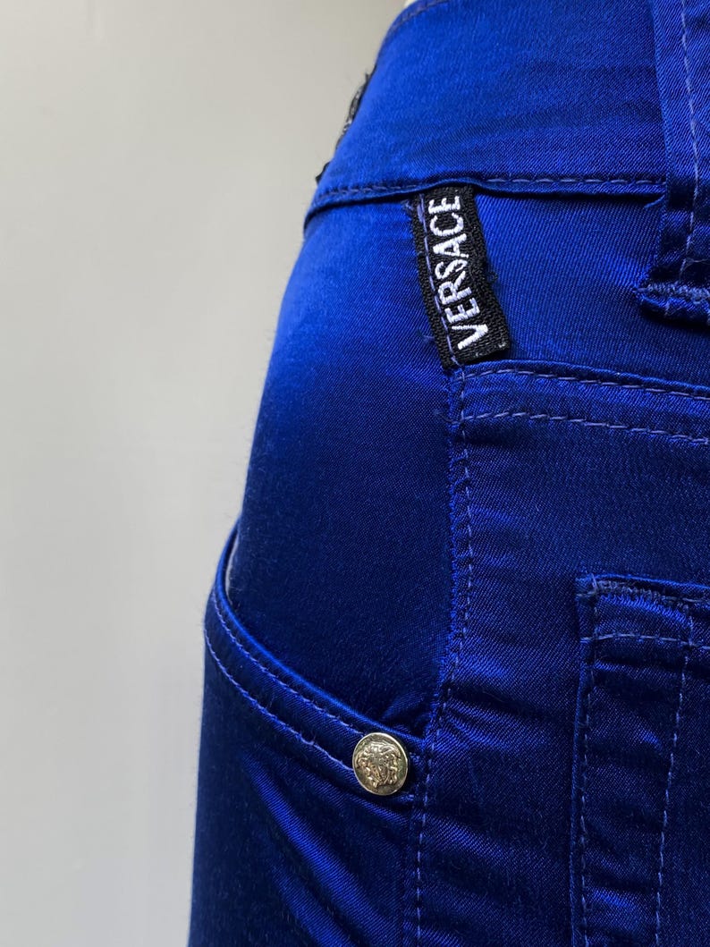 May include: Close-up of a pair of royal blue pants. The pants feature a small pocket, a silver button, and the "VERSACE" label. The fabric has a satin sheen, and the stitching is visible.