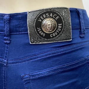 May include: Close-up of blue jeans with a Versace Jeans Couture label. The label is rectangular with a circular emblem featuring a Medusa head. The jeans have visible stitching and a back pocket.