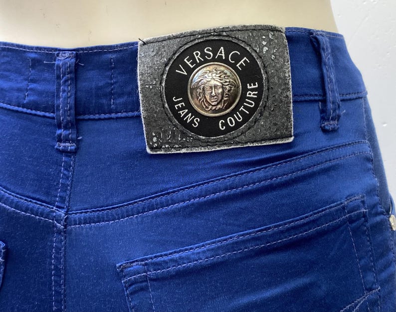 May include: Close-up of blue jeans with a Versace Jeans Couture label. The label is rectangular with a circular emblem featuring a Medusa head. The jeans have visible stitching and a back pocket.