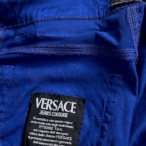 May include: Close-up of a vibrant blue garment with visible stitching details. A black label displays "VERSACE JEANS COUTURE" and Italian text. The garment's fabric appears slightly textured.