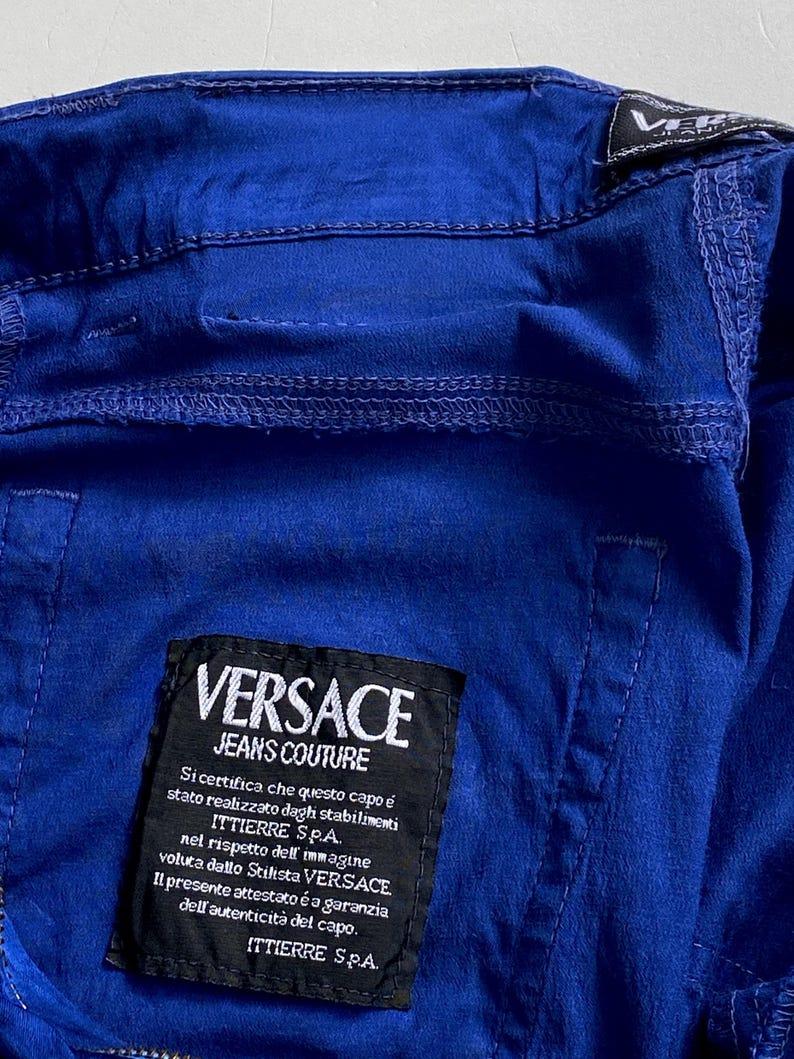 May include: Close-up of a vibrant blue garment with visible stitching details. A black label displays "VERSACE JEANS COUTURE" and Italian text. The garment's fabric appears slightly textured.