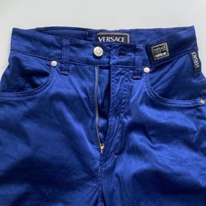 May include: Close-up of blue pants with a zipper and pockets. The pants have the Versace logo on the waistband and a small tag. The pants are made of a shiny material.