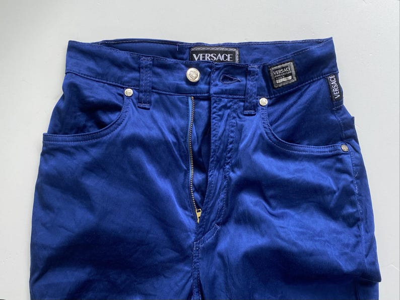 May include: Close-up of blue pants with a zipper and pockets. The pants have the Versace logo on the waistband and a small tag. The pants are made of a shiny material.
