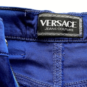 May include: Close-up of blue denim fabric with visible stitching and a black label. The label reads "VERSACE JEANS COUTURE" in white lettering.