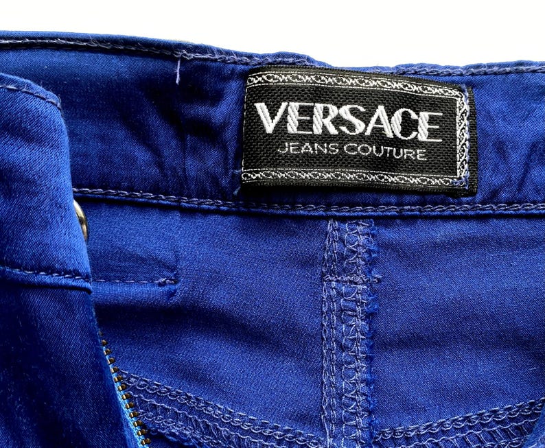 May include: Close-up of blue denim fabric with visible stitching and a black label. The label reads "VERSACE JEANS COUTURE" in white lettering.