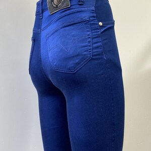 May include: Close-up of blue jeans with a back pocket featuring a stitched design. The jeans have a leather patch with the Versace Jeans Couture logo. The jeans are a solid blue color and have a slim fit.