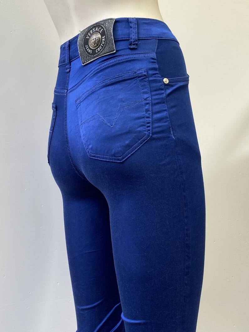 May include: Close-up of blue jeans with a back pocket featuring a stitched design. The jeans have a leather patch with the Versace Jeans Couture logo. The jeans are a solid blue color and have a slim fit.
