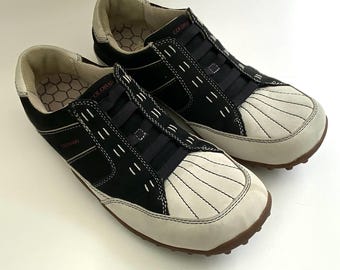 May include: Pair of black and off-white slip-on sneakers. The sneakers have a rounded toe with a fan-like design, black elastic bands across the top, and the word "COLORADO" in pink on the side. The soles are brown.