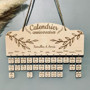 Peut inclure: Un calendrier en bois avec une finition brun clair et une inscription blanche qui dit "Calendrier anniversaires Familles & Amis". Le calendrier comporte 12 mois, chacun avec un petit carré pour chaque jour du mois. Chaque carré porte un nom et une date.