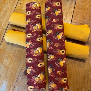 May include: Two red fabric placemats with a pattern of orange pumpkins, sunflowers, and fall leaves. The placemats are rolled up and are sitting on top of four yellow fabric placemats.