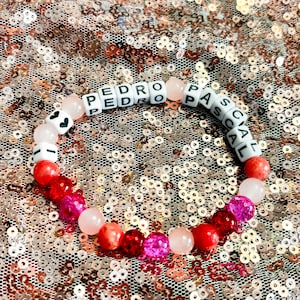 May include: A pink and red beaded bracelet with the name "PEDRO PASCAL" spelled out in white letters.