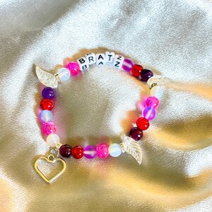 May include: A pink, red, and white beaded bracelet with a silver heart charm and two silver leaf charms. The bracelet spells out "Bratz" in white letters.