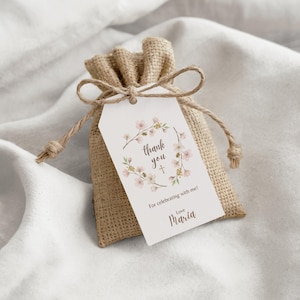 Puede incluir: Una pequeña bolsa de arpillera de color tostado con cordón. Lleva atada una etiqueta rectangular blanca con una ilustración de corona floral, una pequeña cruz y el texto impreso: thank you, For celebrating with me!, Love Maria.