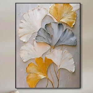 Può includere: Un'opera d'arte incorniciata con foglie di ginkgo stilizzate in bianco, grigio e oro. Le foglie hanno un aspetto strutturato e sono disposte su uno sfondo grigio chiaro. L'opera d'arte è sopra una panca di legno con cuscini e cuscini beige.