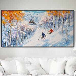 Puede incluir: Pintura al óleo de una pista de esquí nevada con dos esquiadores en rojo y azul. La obra presenta un telesilla y árboles con follaje naranja y blanco. La pintura está enmarcada en negro y cuelga sobre un sofá blanco.