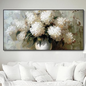 May include: An oil painting of white flowers in a white vase. The flowers are full and lush, with green leaves. The painting is in a black frame and hangs above a white sofa. The background has a textured, neutral color.