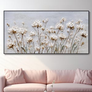 Puede incluir: Pintura floral enmarcada con flores blancas y beige texturizadas. La obra de arte está sobre un fondo gris claro y enmarcada en negro. La pintura se exhibe sobre un sofá rosa en un salón.