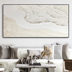 May include: A white abstract painting with a textured surface. The painting is framed in a black frame and is hanging on a white wall above a white couch with brown throw pillows.
