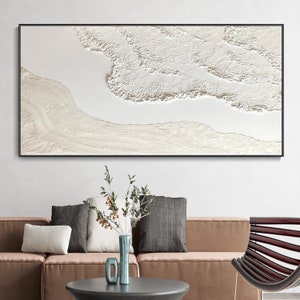 May include: A white abstract painting with a textured surface, framed in black, hanging on a white wall above a brown sofa with pillows.