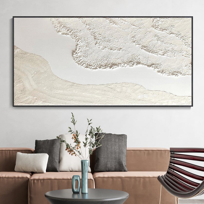 May include: A white abstract painting with a textured surface, framed in black, hanging on a white wall above a brown sofa with pillows.