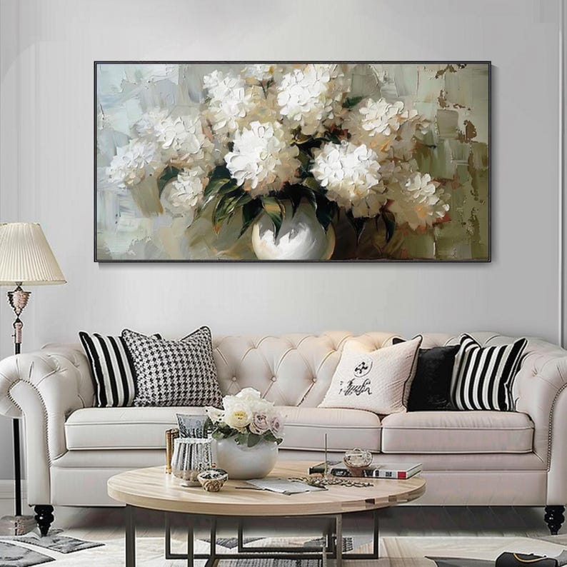 May include: An oil painting of white flowers in a white vase. The artwork is in a black frame and hangs above a white sofa with decorative pillows. The painting has a textured, impressionistic style.