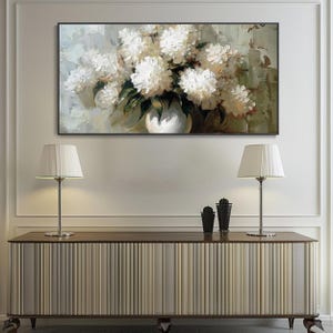 May include: An oil painting depicts a bouquet of white flowers in a white vase. The artwork is set against a muted background with green and beige tones. The painting is framed and hangs above a wooden cabinet with two lamps.