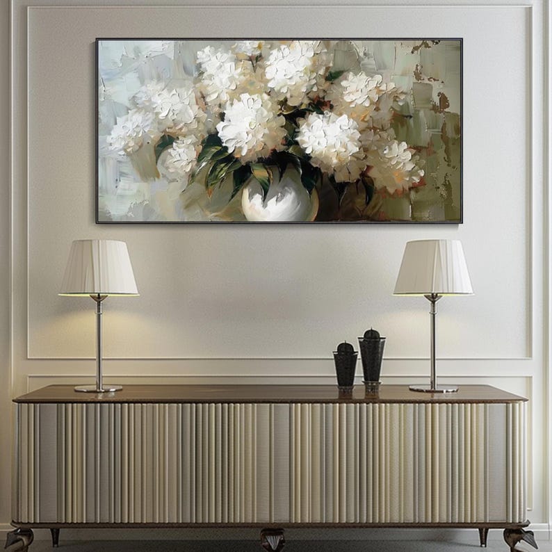 May include: An oil painting depicts a bouquet of white flowers in a white vase. The artwork is set against a muted background with green and beige tones. The painting is framed and hangs above a wooden cabinet with two lamps.