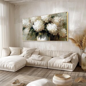 May include: A large, rectangular oil painting featuring a bouquet of white flowers in a white vase. The artwork has a gold frame and hangs above a cream-colored sectional sofa. The painting's background is a muted green and beige.
