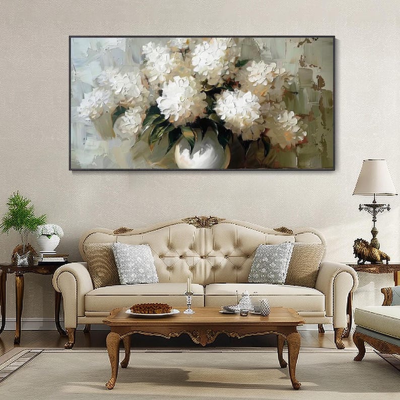 May include: A framed oil painting depicts a bouquet of white flowers in a white vase. The artwork is set against a neutral background with visible brushstrokes. The painting hangs above a cream-colored sofa in a living room setting.