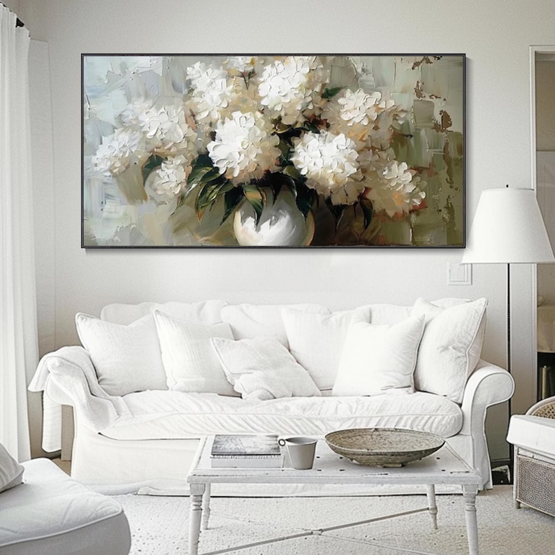 May include: An oil painting of white flowers in a white vase. The flowers are full and lush, with green leaves. The painting is in a black frame and hangs above a white sofa. The background has a textured, neutral color.