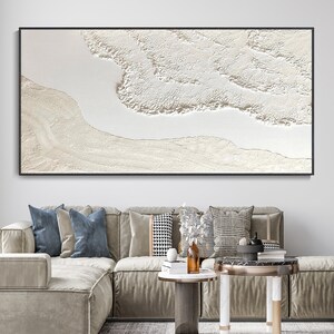 May include: A large abstract painting with a white background and a textured, raised design in shades of white and cream. The painting is framed in a black frame and is hanging on a white wall.