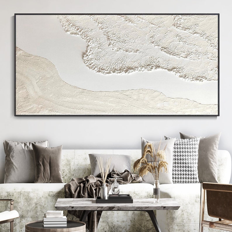 May include: A white abstract painting with a textured surface. The painting is framed in a black frame and is hanging on a white wall above a white couch with brown throw pillows.