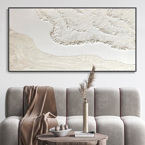 May include: A white abstract painting with a textured, layered design. The painting is framed in black and hangs on a white wall above a light brown couch.