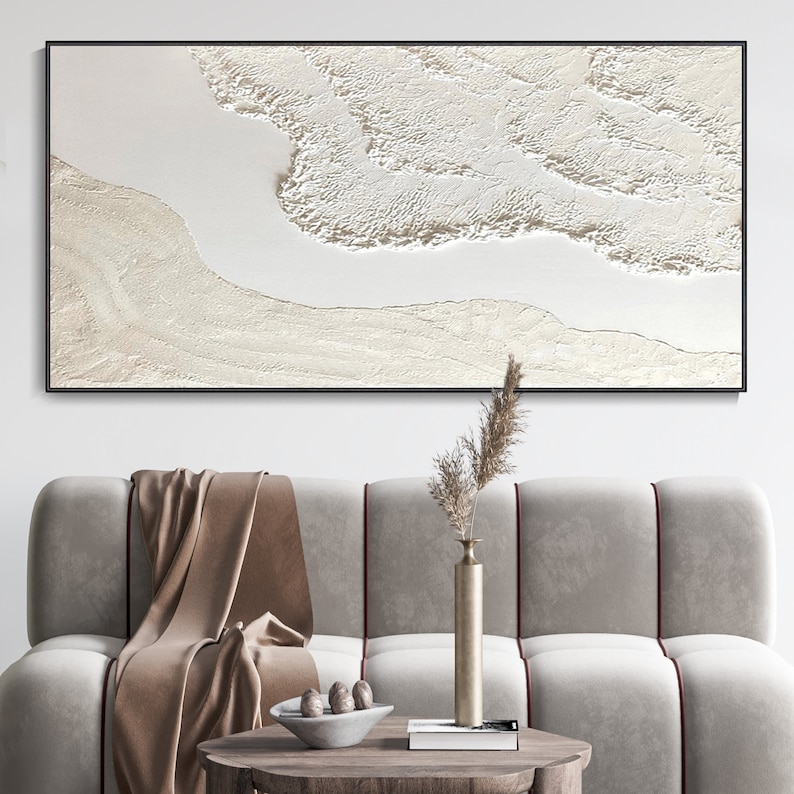 May include: A white abstract painting with a textured, layered design. The painting is framed in black and hangs on a white wall above a light brown couch.