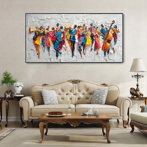 K&ouml;nnte beinhalten: Gerahmtes &Ouml;lgem&auml;lde, das eine lebendige Gruppe von Musikern in Bewegung zeigt. Das Kunstwerk zeigt einen strukturierten wei&szlig;en Hintergrund mit Figuren in leuchtenden Farben wie Rot, Blau, Gelb und Lila, die Instrumente spielen. Das Gem&auml;lde h&auml;ngt &uuml;ber einem cremefarbenen Sofa.