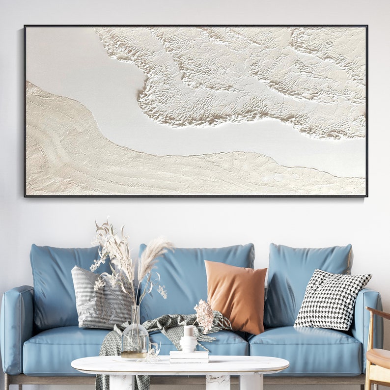 May include: A white abstract painting with a textured surface. The painting is framed in black and hangs on a white wall above a blue sofa.