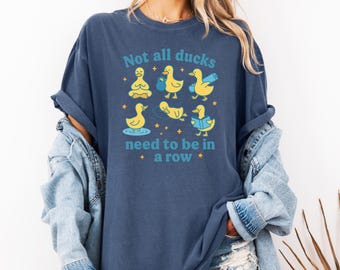 May include: A navy blue t-shirt with the text "Not all ducks need to be in a row" and illustrations of yellow ducks in various poses. The shirt is made of a soft material and has rolled-up sleeves.