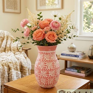 May include: A ceramic vase with a pink and white fan-shaped pattern containing a floral arrangement of peach roses, pink ranunculus, white cosmos, and green eucalyptus stems. The vase has a flared rim and a rounded body with a glossy finish.