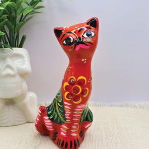 May include: A red ceramic cat figurine with black accents and floral designs. The cat has a whimsical expression and is decorated with yellow, pink, and green details. The figurine is marked with the number 1.