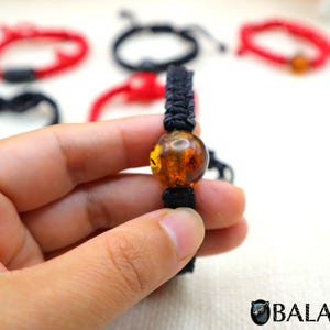 May include: A black braided bracelet with a large, round amber bead. Several other bracelets in red and black are visible in the background. The amber bead is a warm, translucent orange color. The bracelet is held in a person's hand.